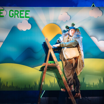 A person wearing a troll costume stood on a wooden ladder. A mountain landscape painting in the background. The top left of the painting read THE GREE