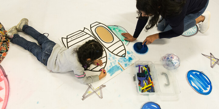 A young person and woman coloring a paper with rocket outlines on it. Colouring pencils placed on the floor.