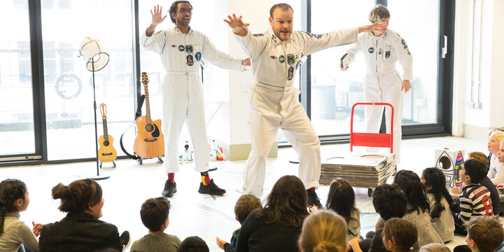 Three people dressed in white astronaut costume. A group of children sat watching them.