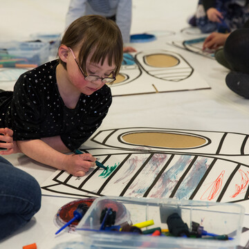 A young person wearing glasses, coloring inside an outline of a rocket.