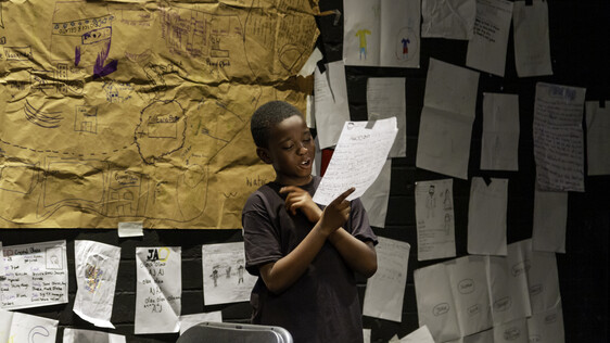 Boy reads from a page - behind him is a wall with lots of pages pinned to it.
