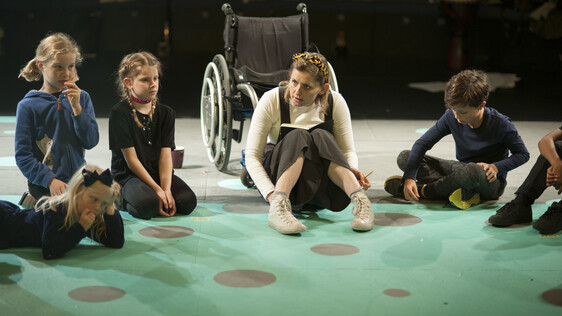 Workshop leader sat in a circle with children, there is a wheelchair behind her.